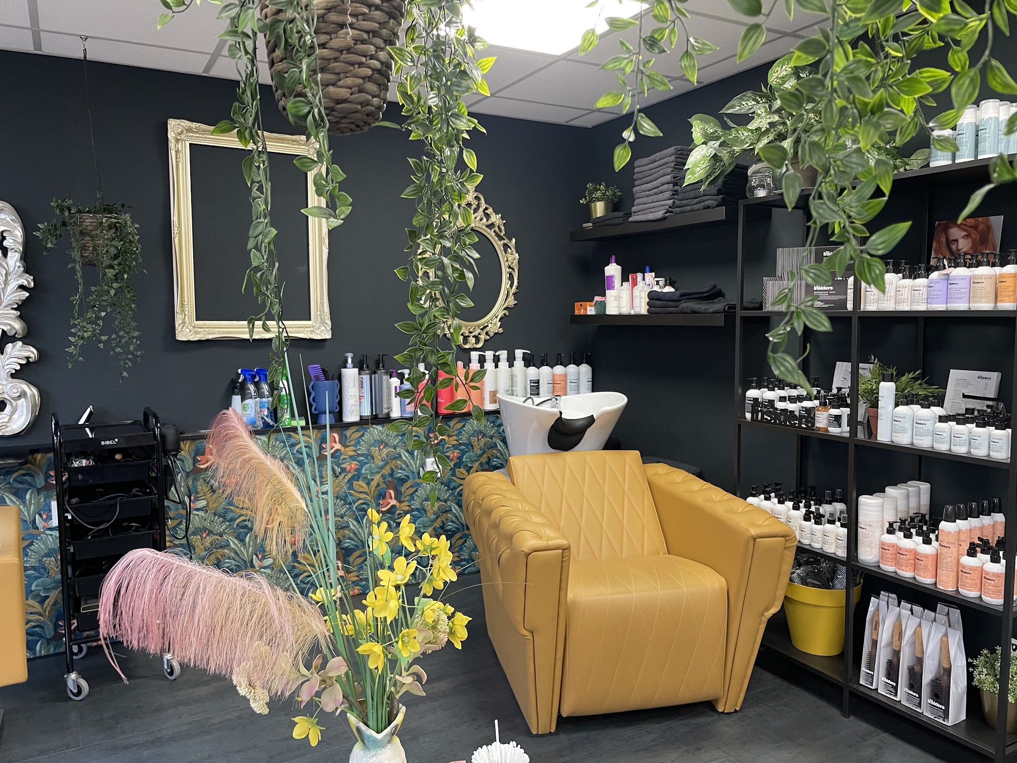 De salon: donkere muur met gouden lijsten, gele stoelen, planten en schappen met producten