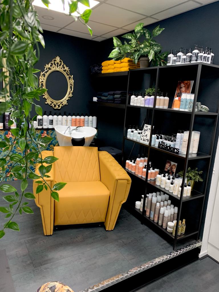 Hoek van de salon met een gele wasstoel, een gouden ovale lijst en een zwarte kast met haarproducten